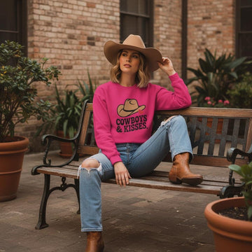 Woman wearing Cowboys And Kisses Sweatshirt With Cowboy Hat Heart Graphic, sitting on a bench in jeans and brown boots.